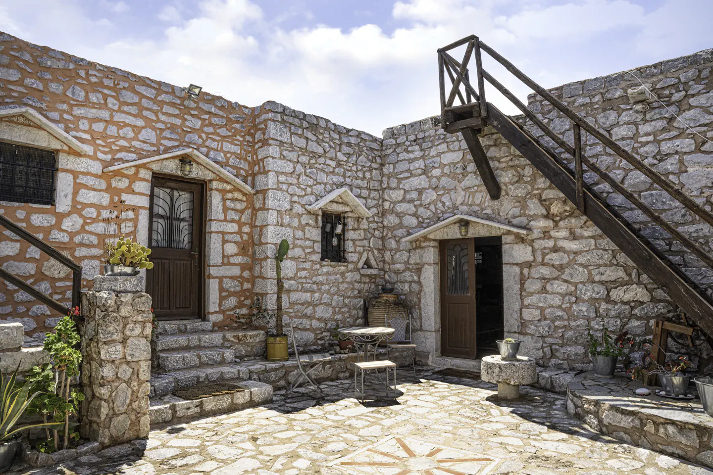 Stone courtyard with two brown doors, windows, and a wooden staircase. Table and chairs sit on the patterned stone floor.