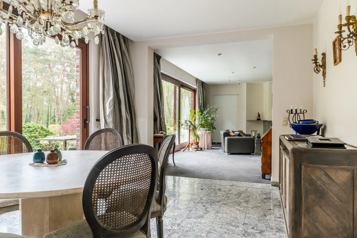 A dining room with a marble floor, a round table with chairs, and a view of the garden through a large window.