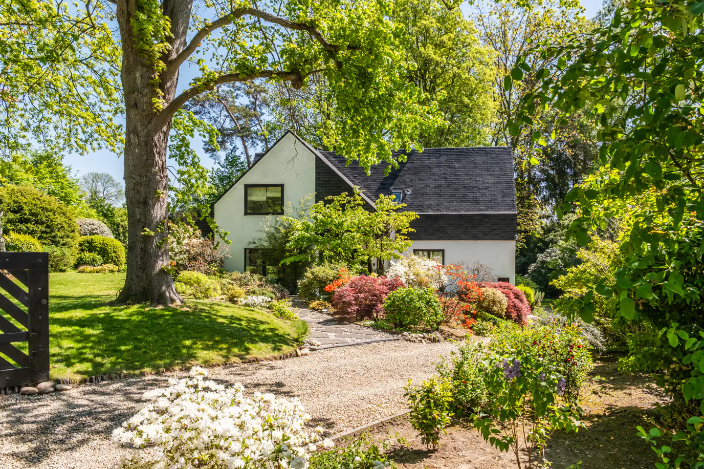 A white house with a black roof surrounded by lush green trees and colorful flowers.