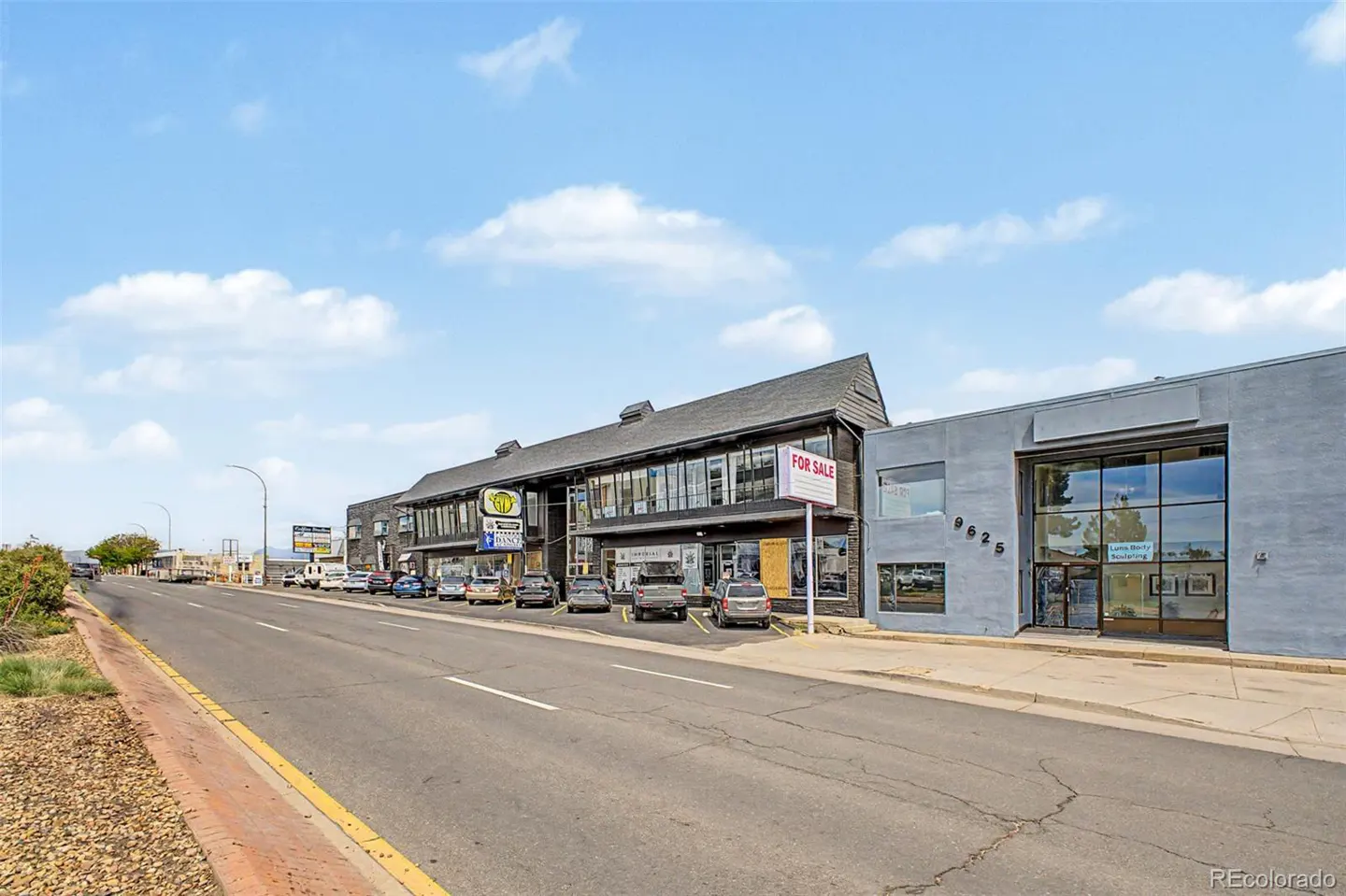 Commercial street view of a multi-story building with a For Sale sign, adjacent to a grey building under a blue sky.