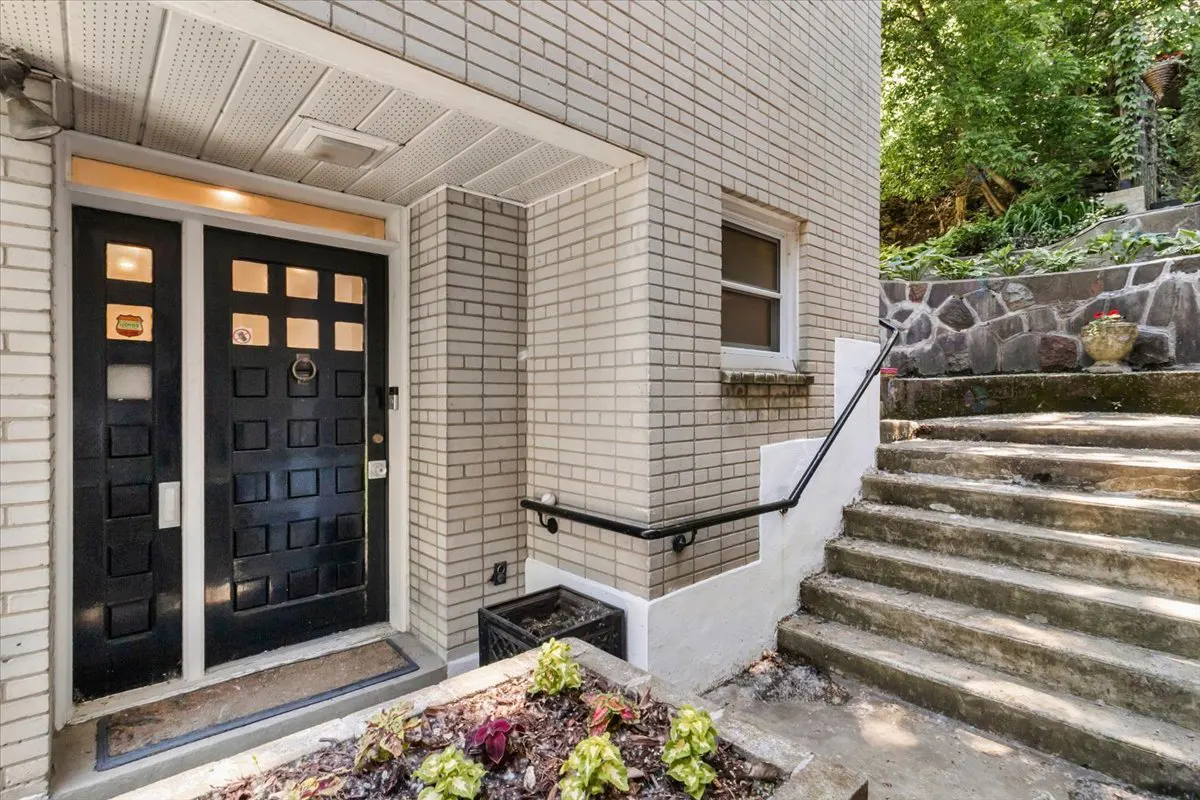 Black front door with square panels next to light brick exterior, a handrail, and stone steps leading to a garden.