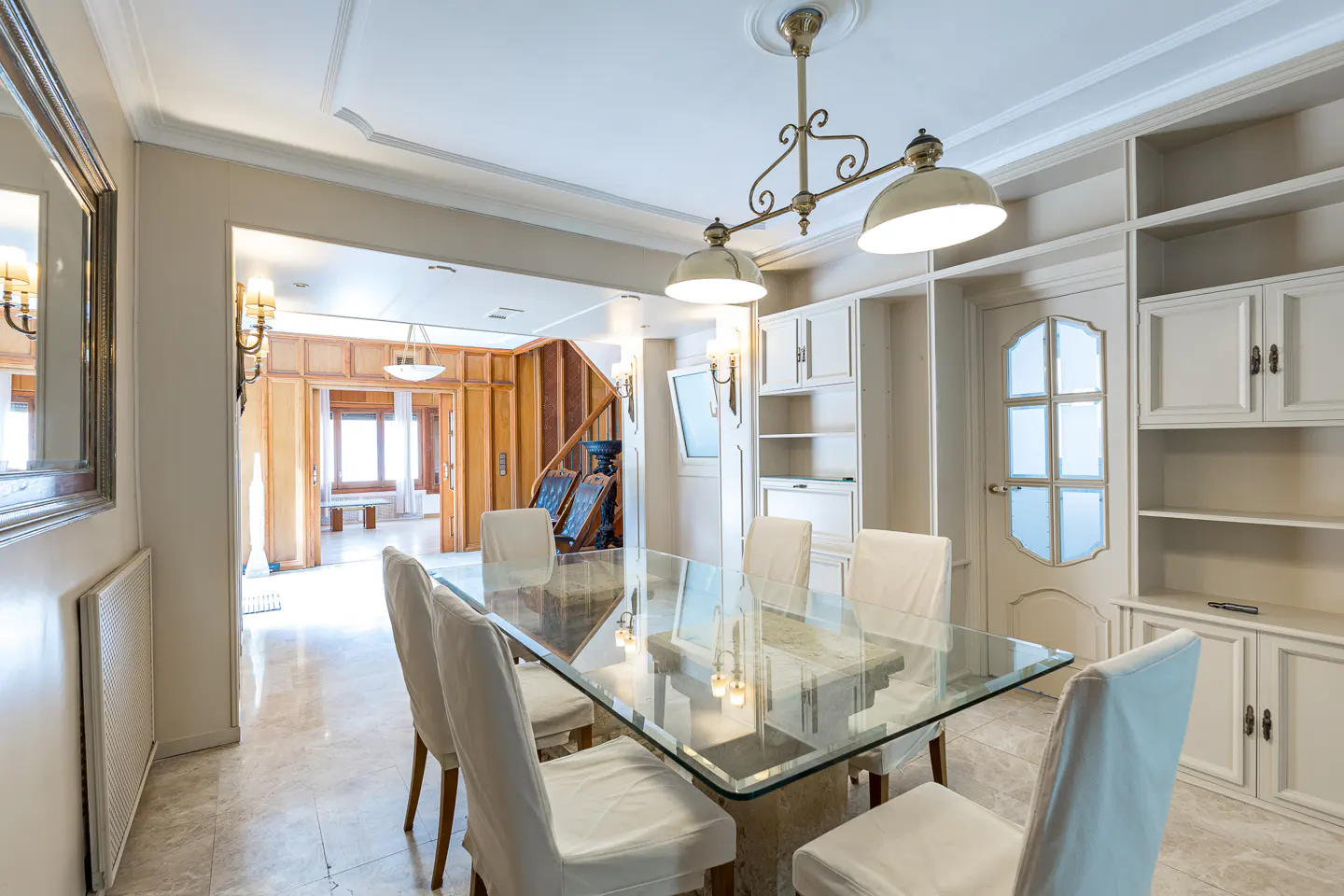 A dining room with a glass table, white chairs, and built-in cabinets. A chandelier hangs above the table.