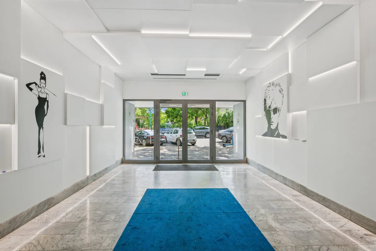 Modern building lobby with white walls, marble floors, and glass doors. Artwork of Audrey Hepburn and Andy Warhol adorn the walls. A blue rug leads to the entrance.