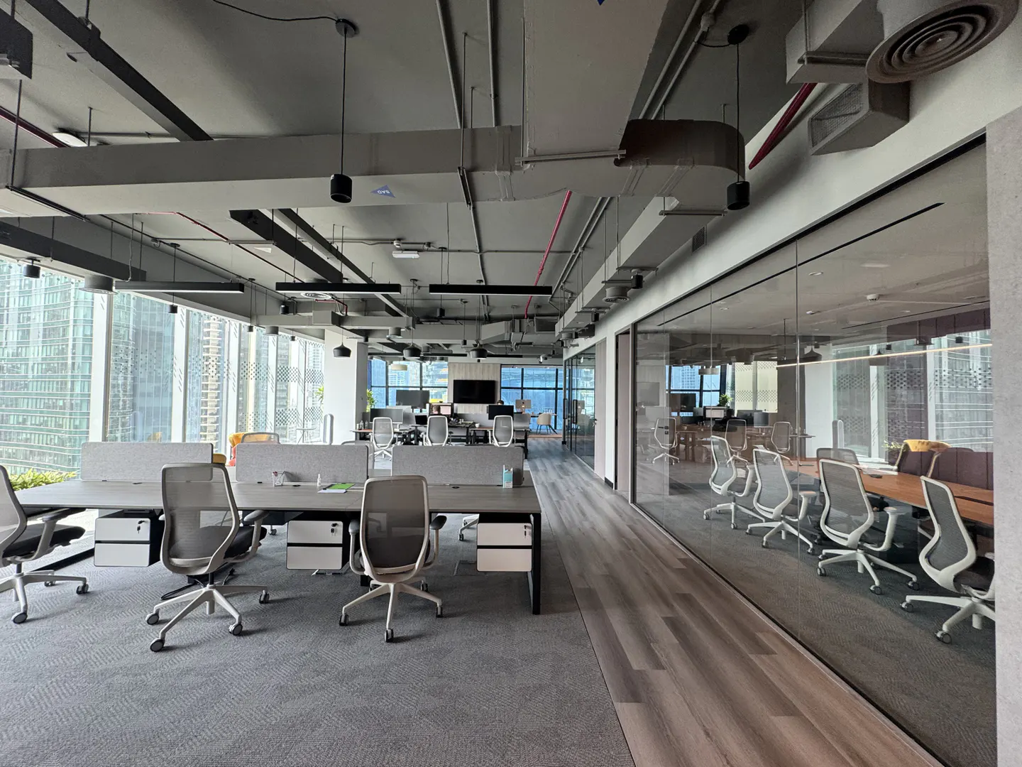 Open office space with grey desks, chairs, and carpet. Large windows offer city views. A glass-walled conference room is visible.