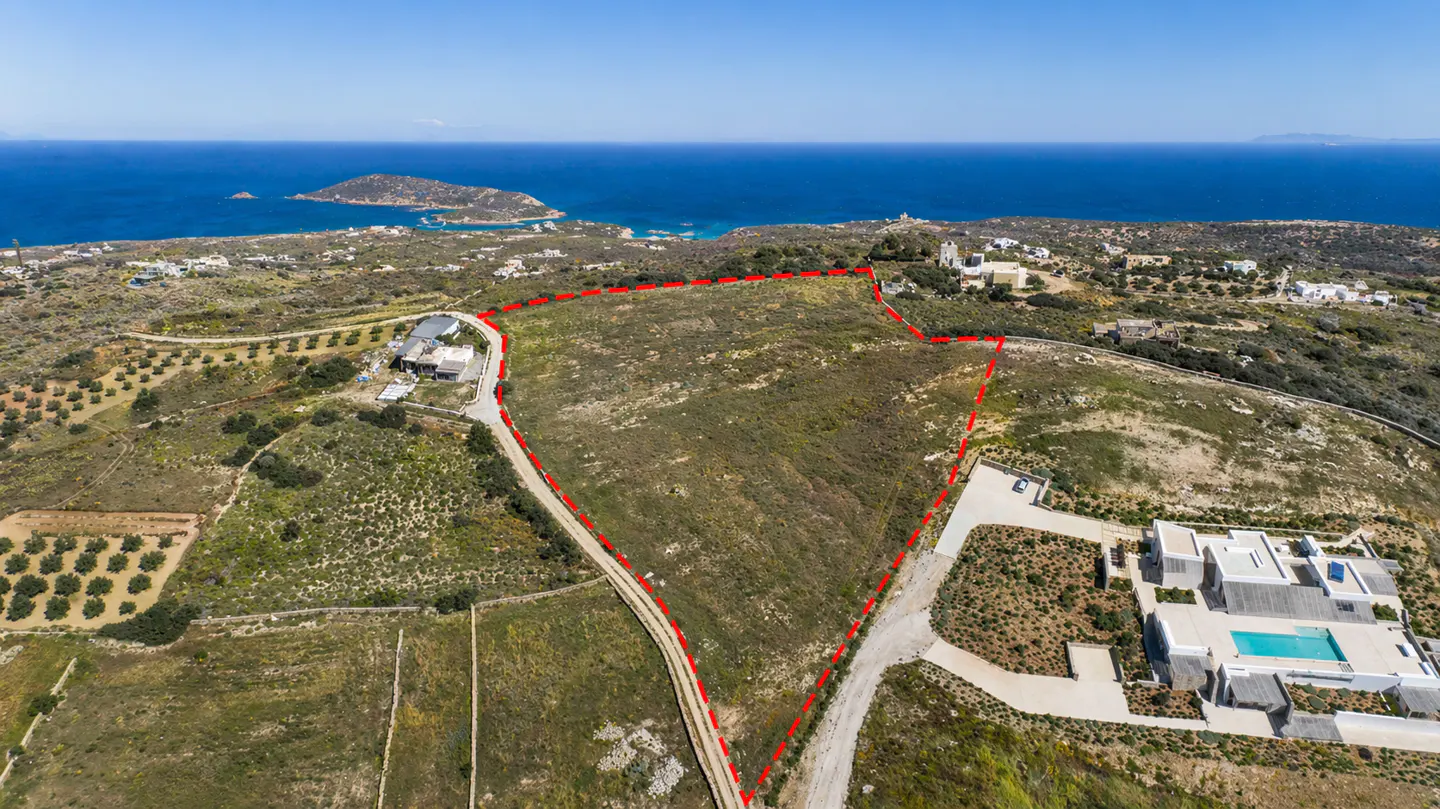 Aerial view of a large, undeveloped plot of land marked with a red dotted line, near the ocean and a modern white house.