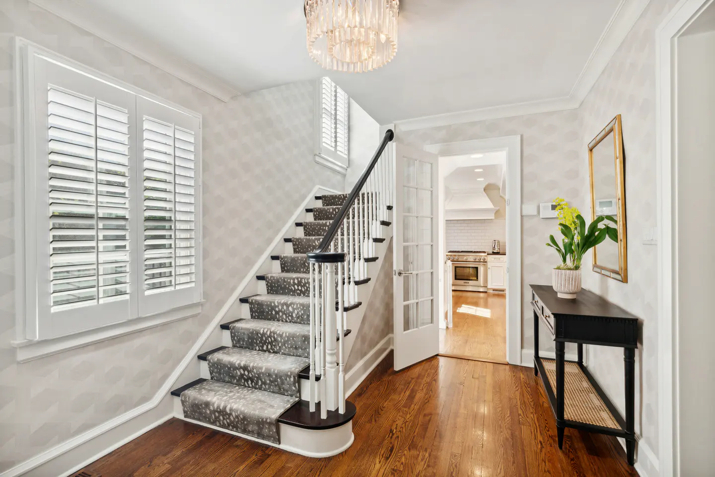 Foyer with hardwood floors, stairs with patterned carpet, and a black console table with a plant. A crystal chandelier hangs from the ceiling.