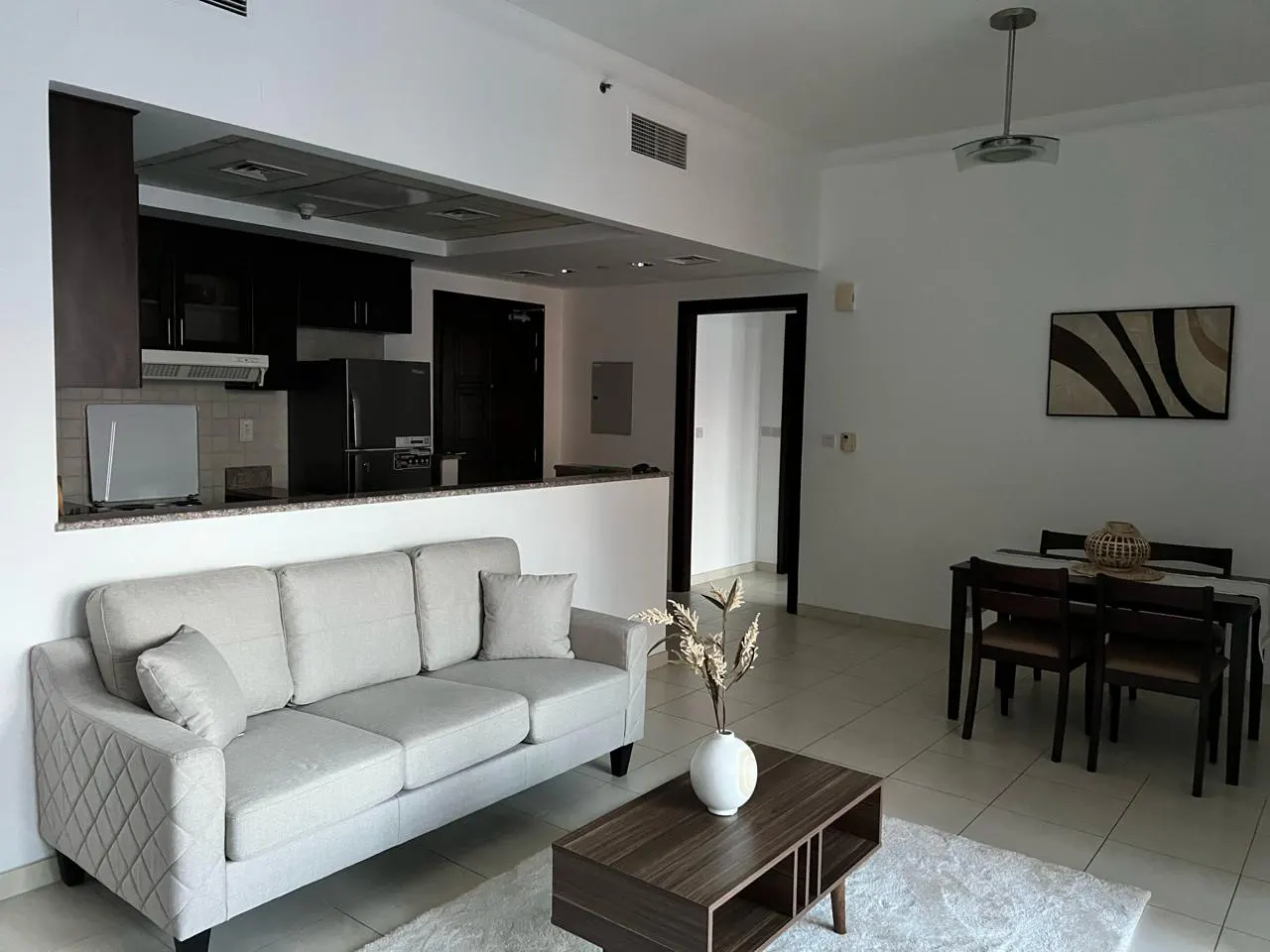 A bright apartment living space with a gray sofa, dark wood coffee table, and dining set. The kitchen is visible in the background.