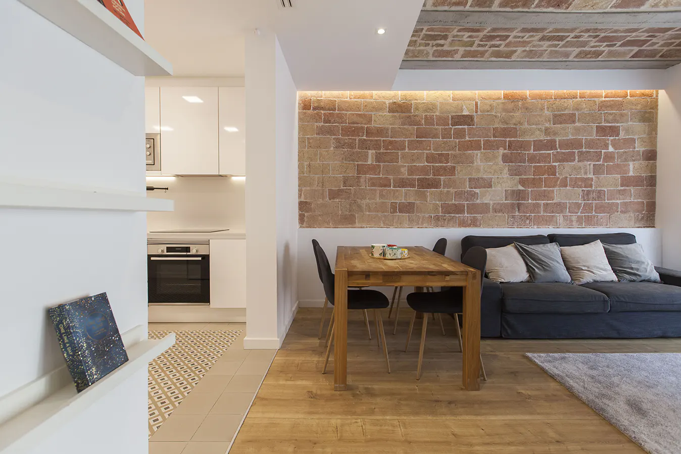 Interior view of a modern apartment with a brick wall, wooden table, gray sofa, and a glimpse of a white kitchen.