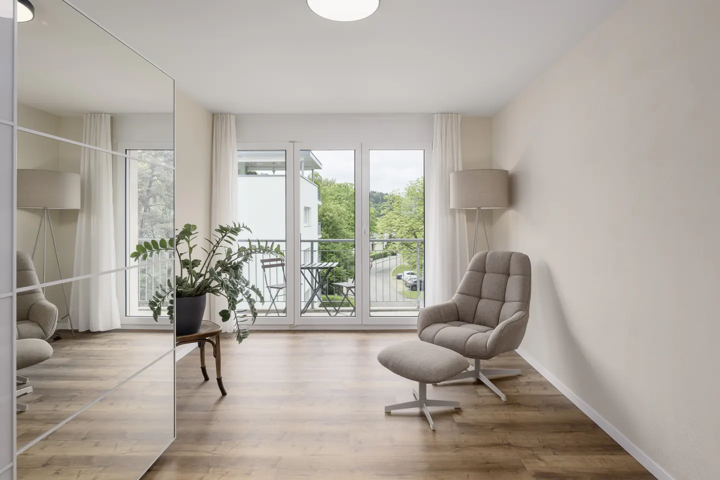 Bright living room with wood floors, beige walls, and a balcony. A gray chair and ottoman sit near a floor lamp. A mirror reflects the room.