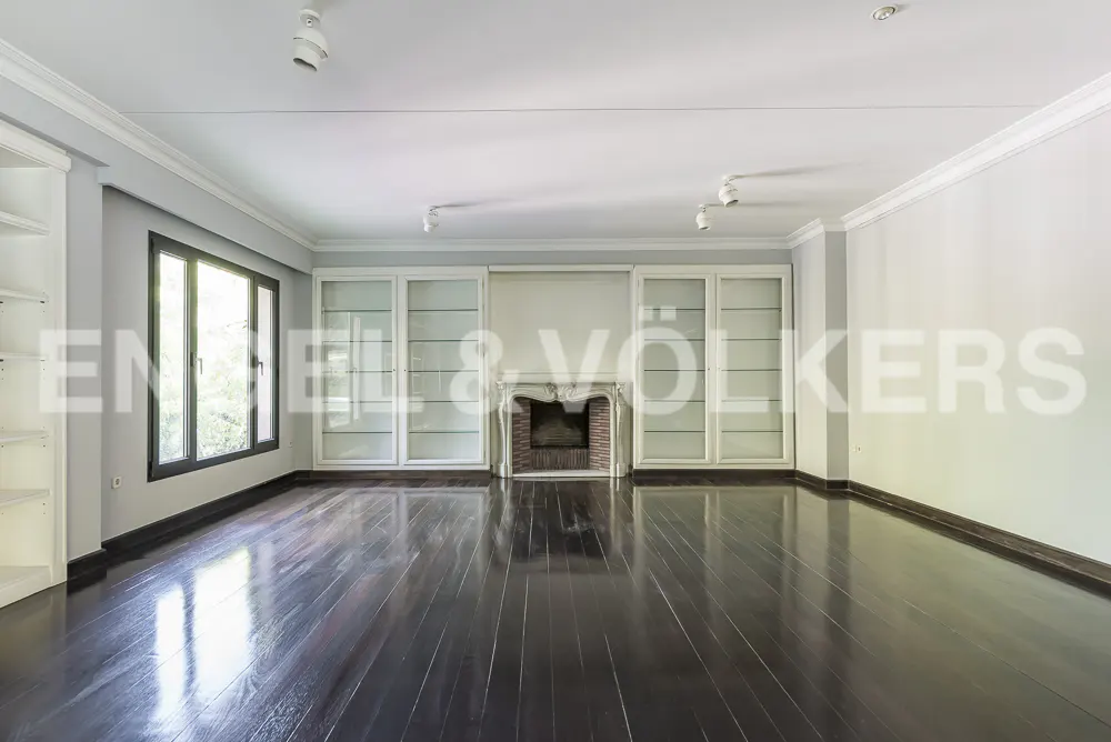A spacious living room with dark wood floors, a fireplace, and built-in white cabinets. A large window lets in natural light.