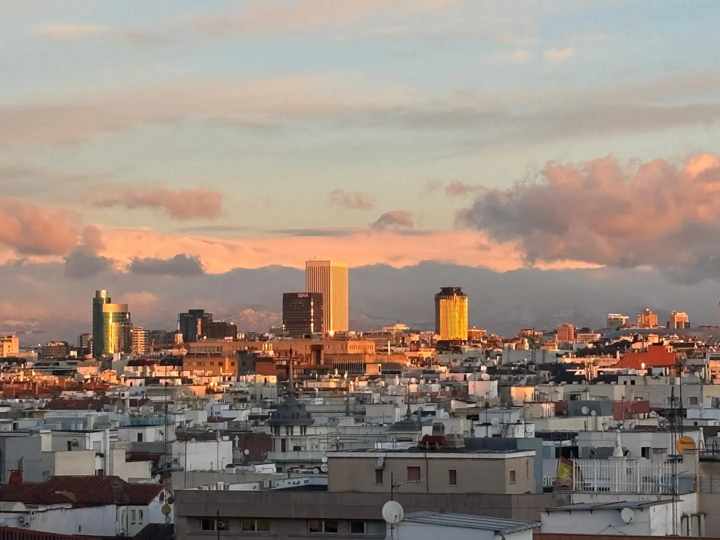 Cityscape view of Madrid, Spain, at sunset. Buildings are white and brown, with skyscrapers in the background. The sky is blue and pink.