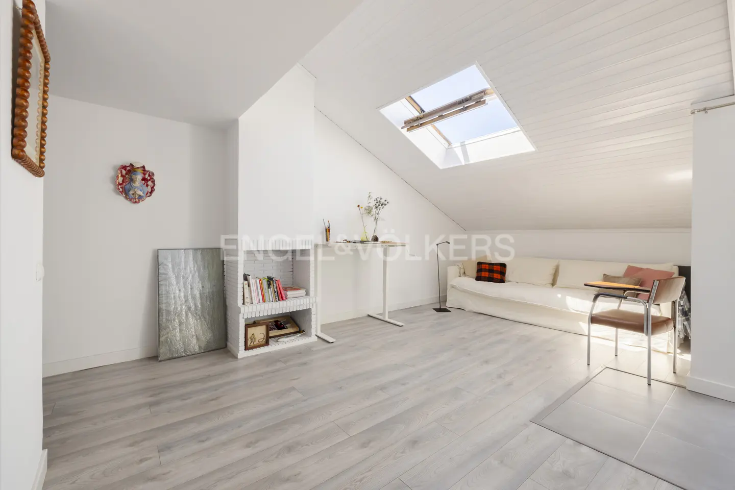 Bright attic living room with white walls, light wood floors, a skylight, a white sofa, and a small desk area.