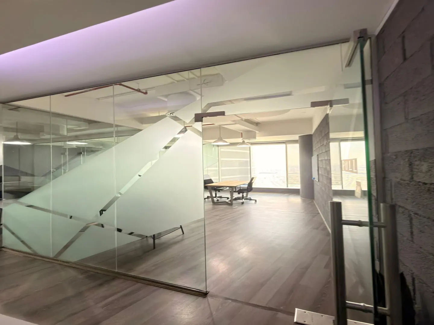 Modern office interior with glass walls, wood-look flooring, and a conference table with chairs in a bright, open space.