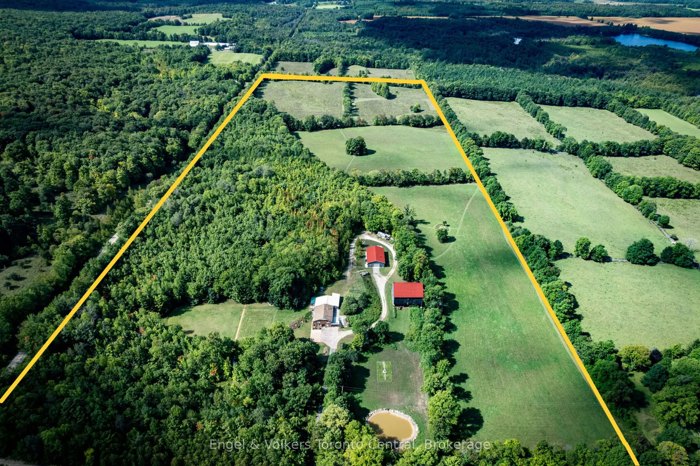 Aerial view of a rural property outlined in yellow, featuring two red-roofed buildings, green fields, and dense forest.