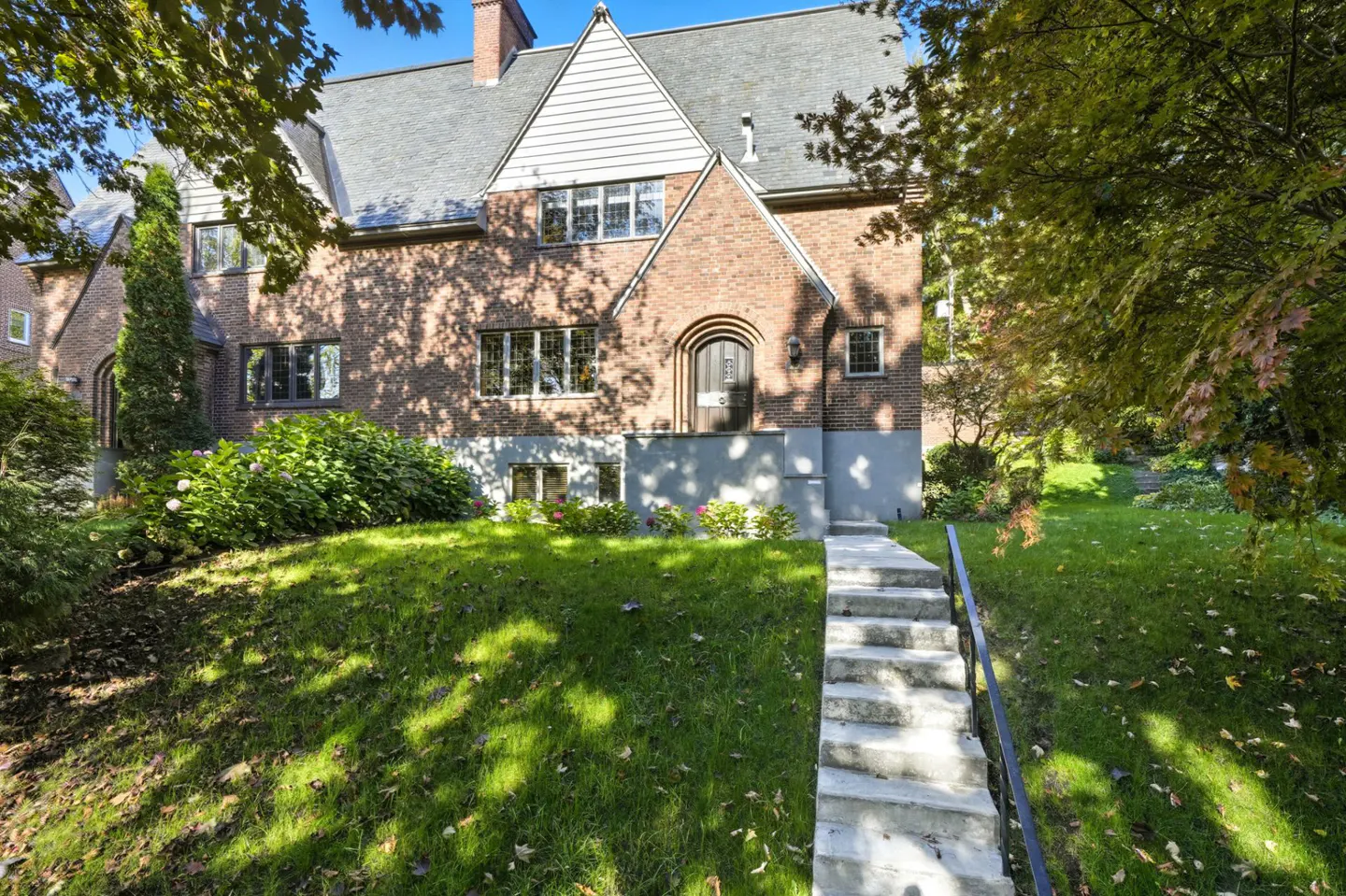 Brick Tudor-style home with gray roof, white gable, and arched doorway. Concrete steps lead up to the house through a green lawn.