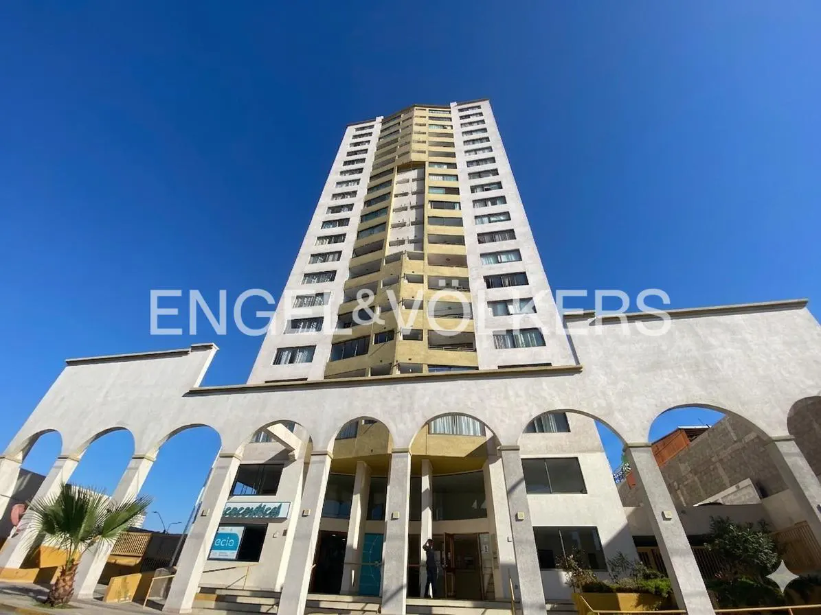 Exterior shot of a tall, modern apartment building with white arches at the entrance under a clear blue sky.