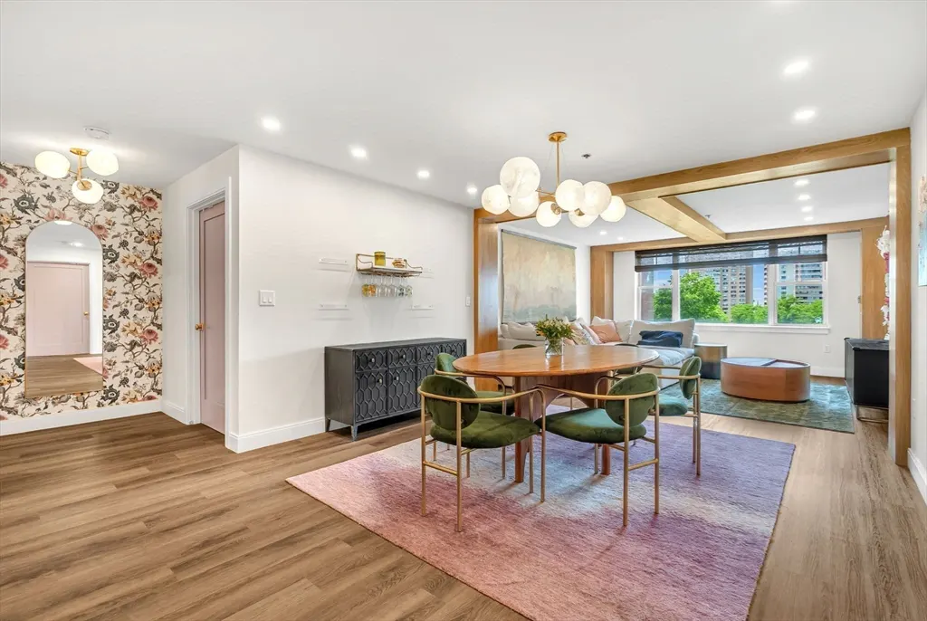 Dining area with a round wooden table, green velvet chairs, pink ombre rug, and a modern chandelier on wood floors.