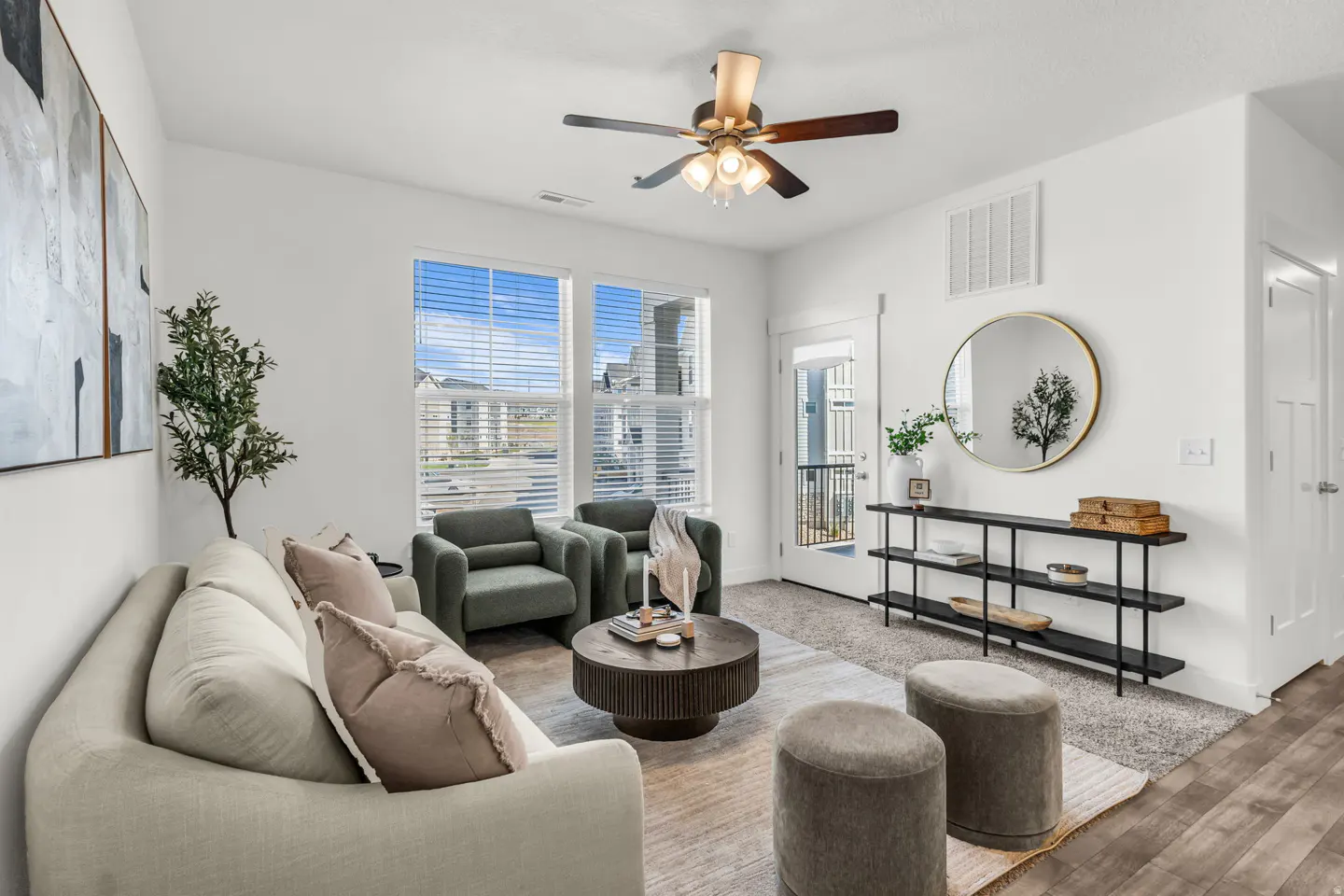 Bright living room with white walls, a beige sofa, two green armchairs, and a round coffee table. A ceiling fan hangs above.