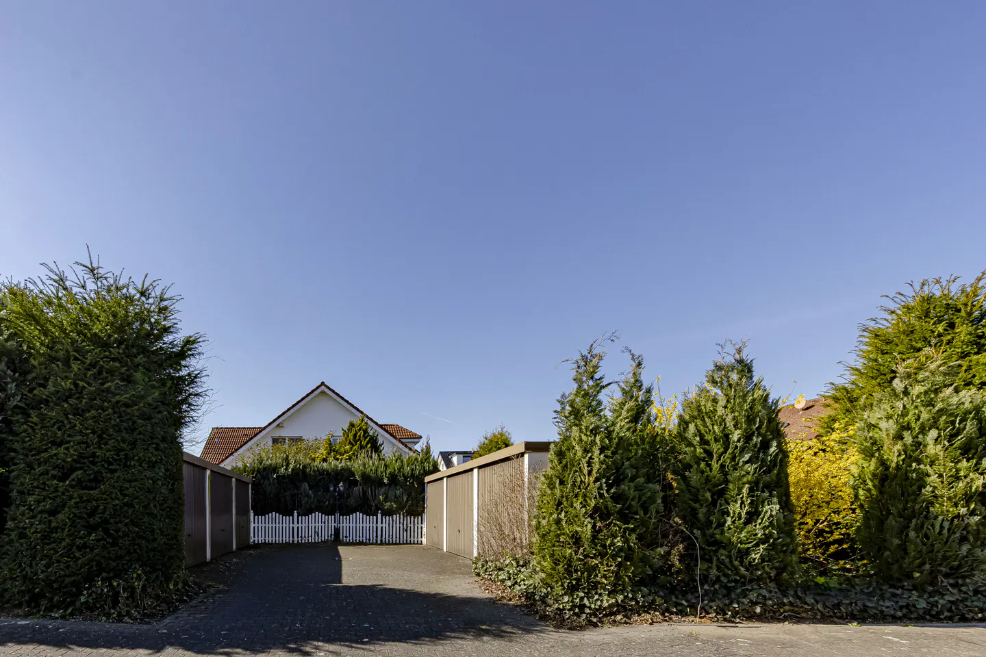 View of a house with a white picket fence and a brown garage, framed by green trees under a clear blue sky.