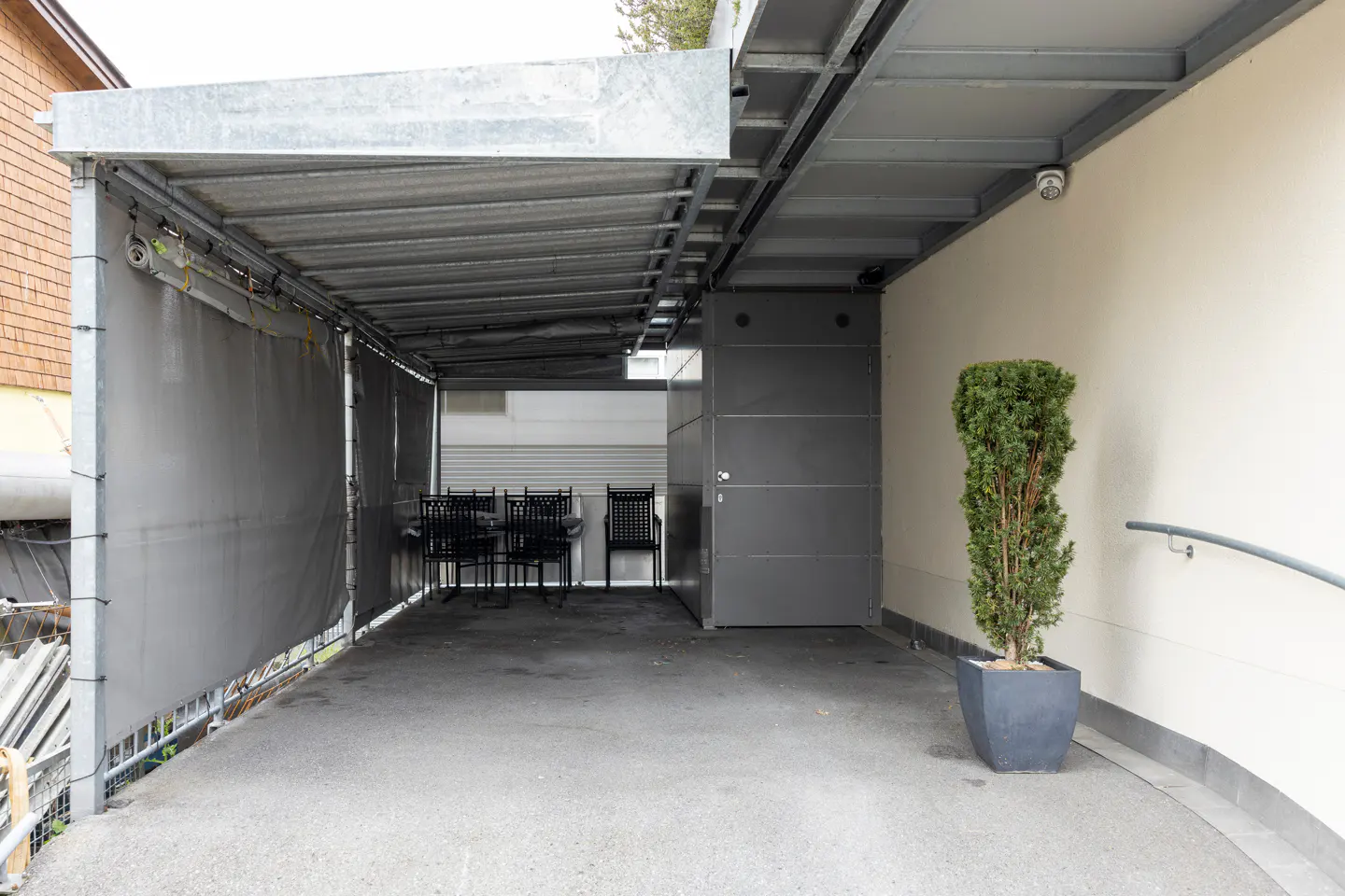 Covered outdoor patio with a metal roof, grey privacy screen, dining set, and a potted evergreen tree against a white wall.