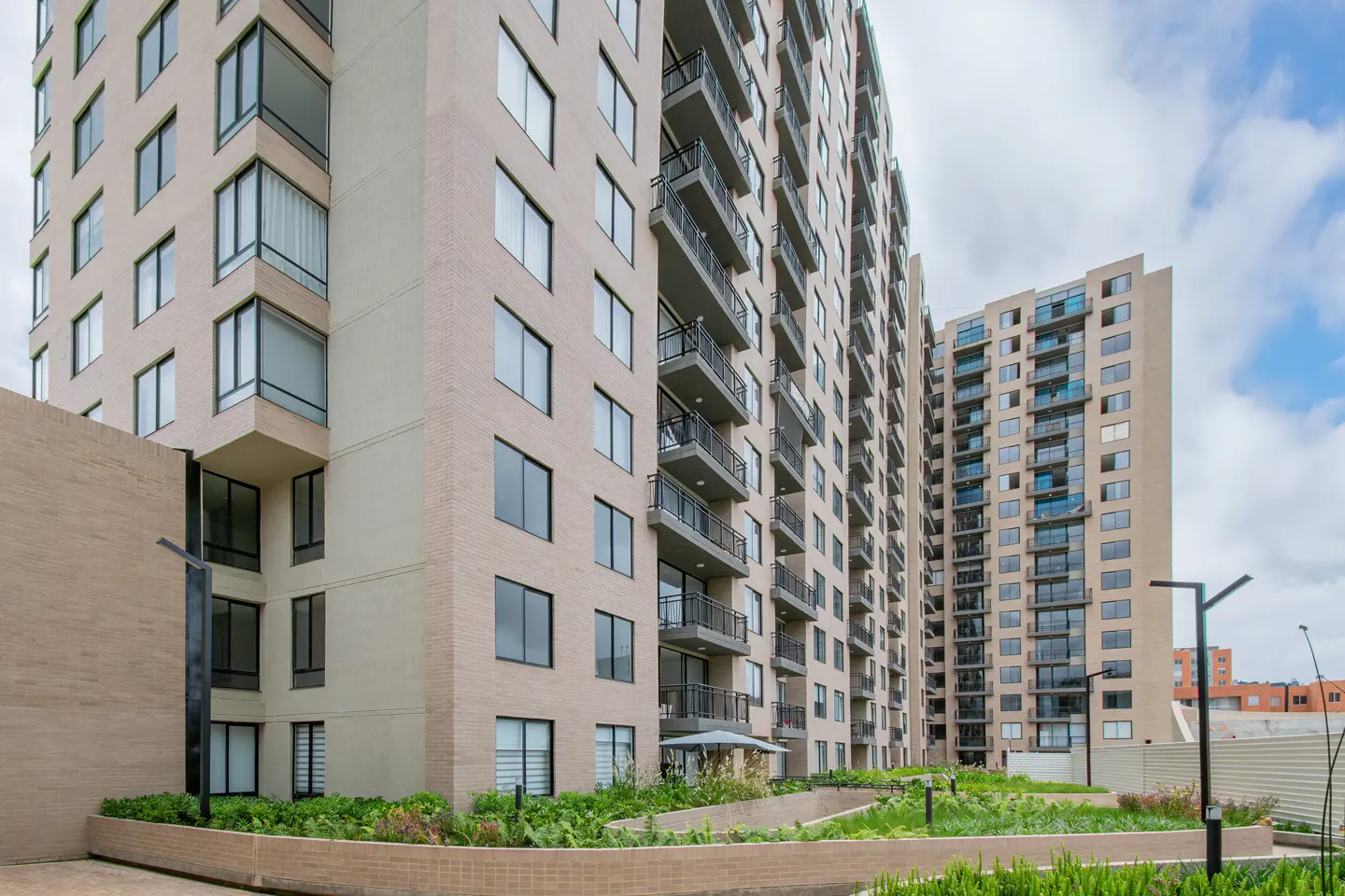 Exterior view of two tall, beige apartment buildings with black balconies and many windows, surrounded by green landscaping.