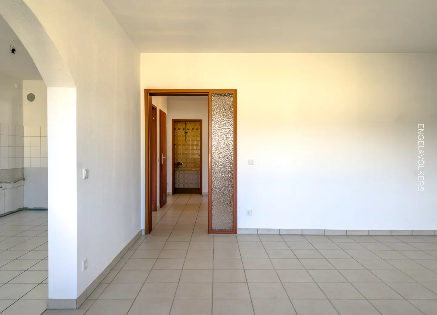 Empty room with white walls and beige tile floor. A doorway leads to a hallway with brown doors and a glass panel. Kitchen visible through an archway.