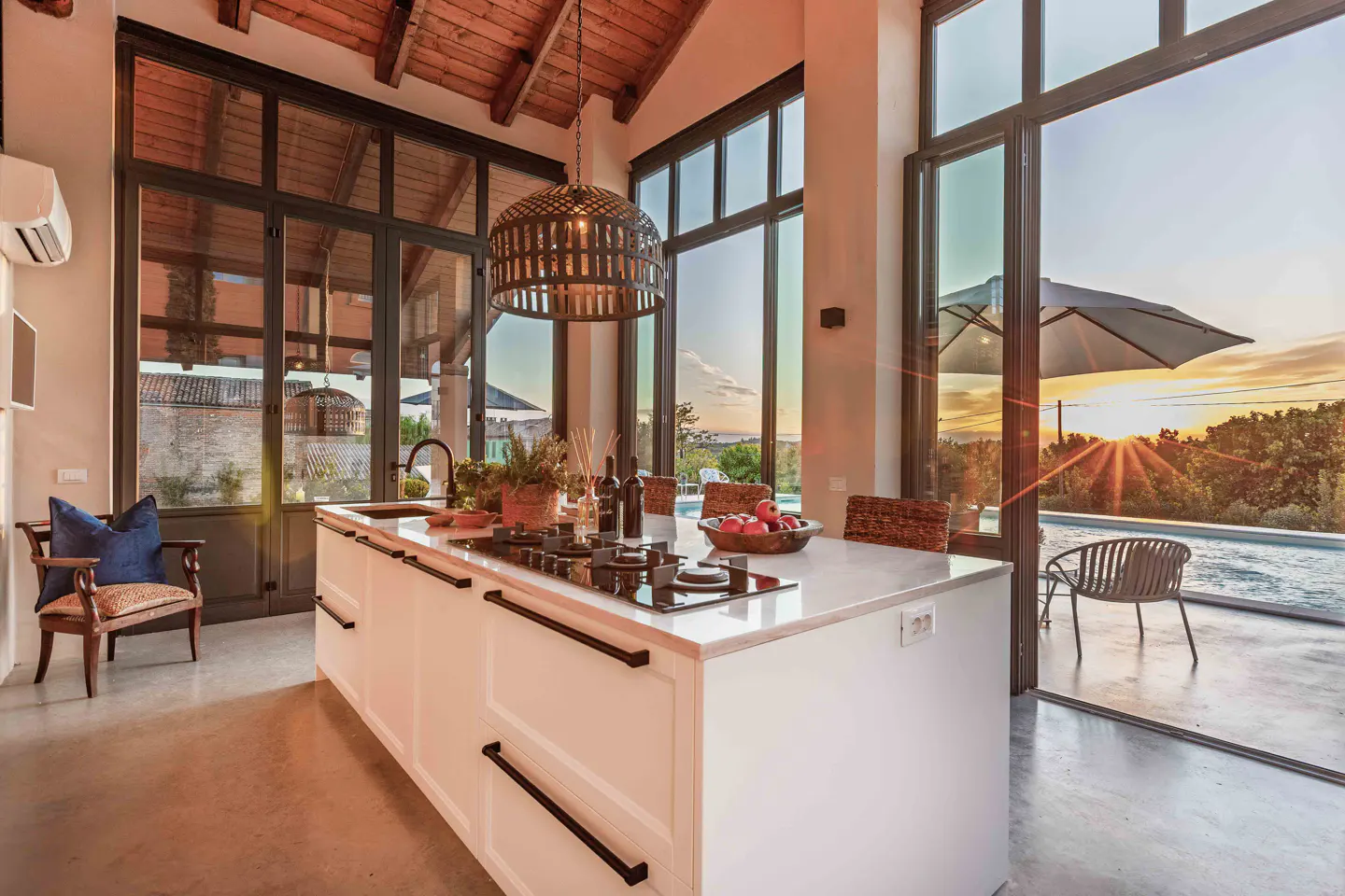 Bright kitchen with white island, black accents, and large windows overlooking a pool at sunset.