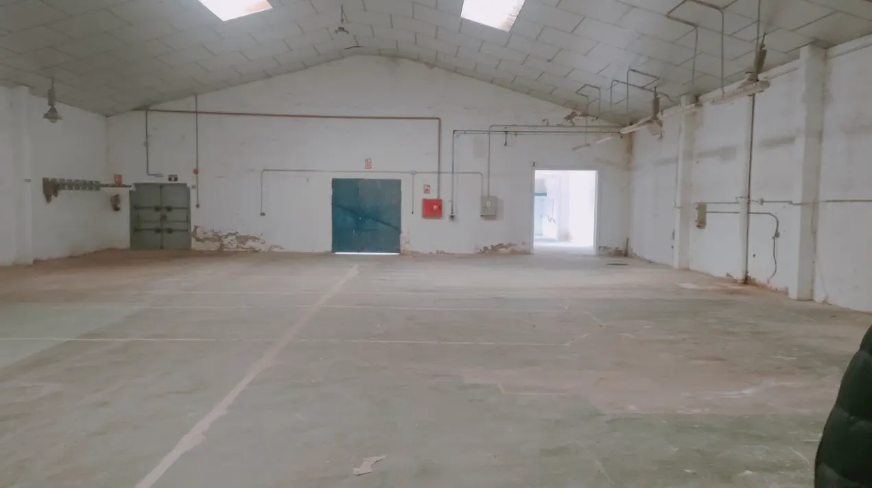A large, empty warehouse with white walls, a concrete floor, and a high ceiling with skylights. There are two blue doors and a red fire extinguisher on the walls.