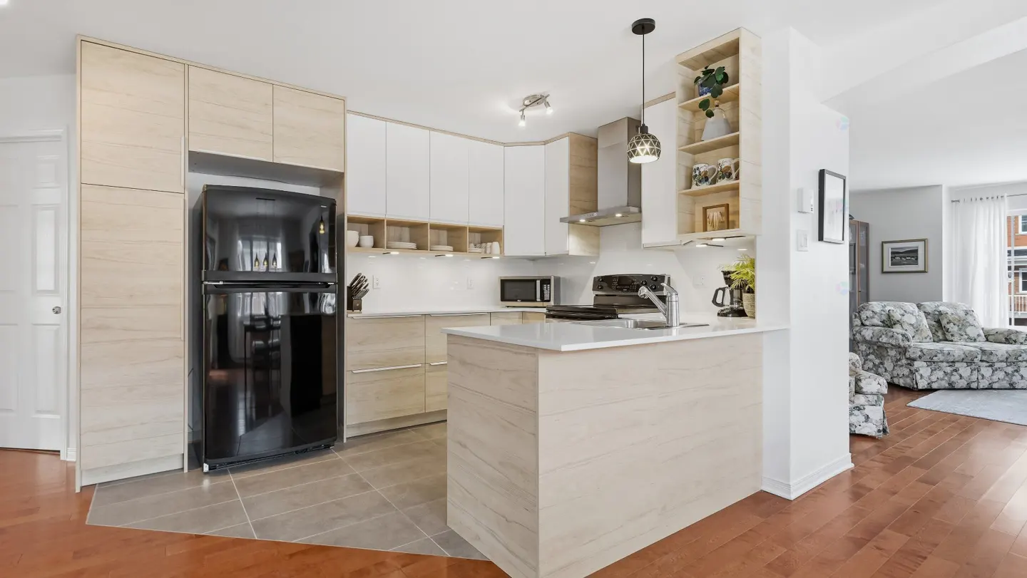 A modern kitchen with light wood cabinets, white countertops, and a black refrigerator. A kitchen island faces a living room with floral sofa.