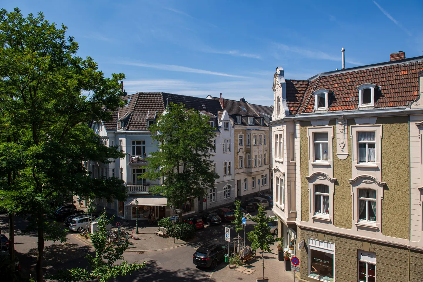 Street view of European-style buildings with red tile roofs, green trees, parked cars, and blue sky.