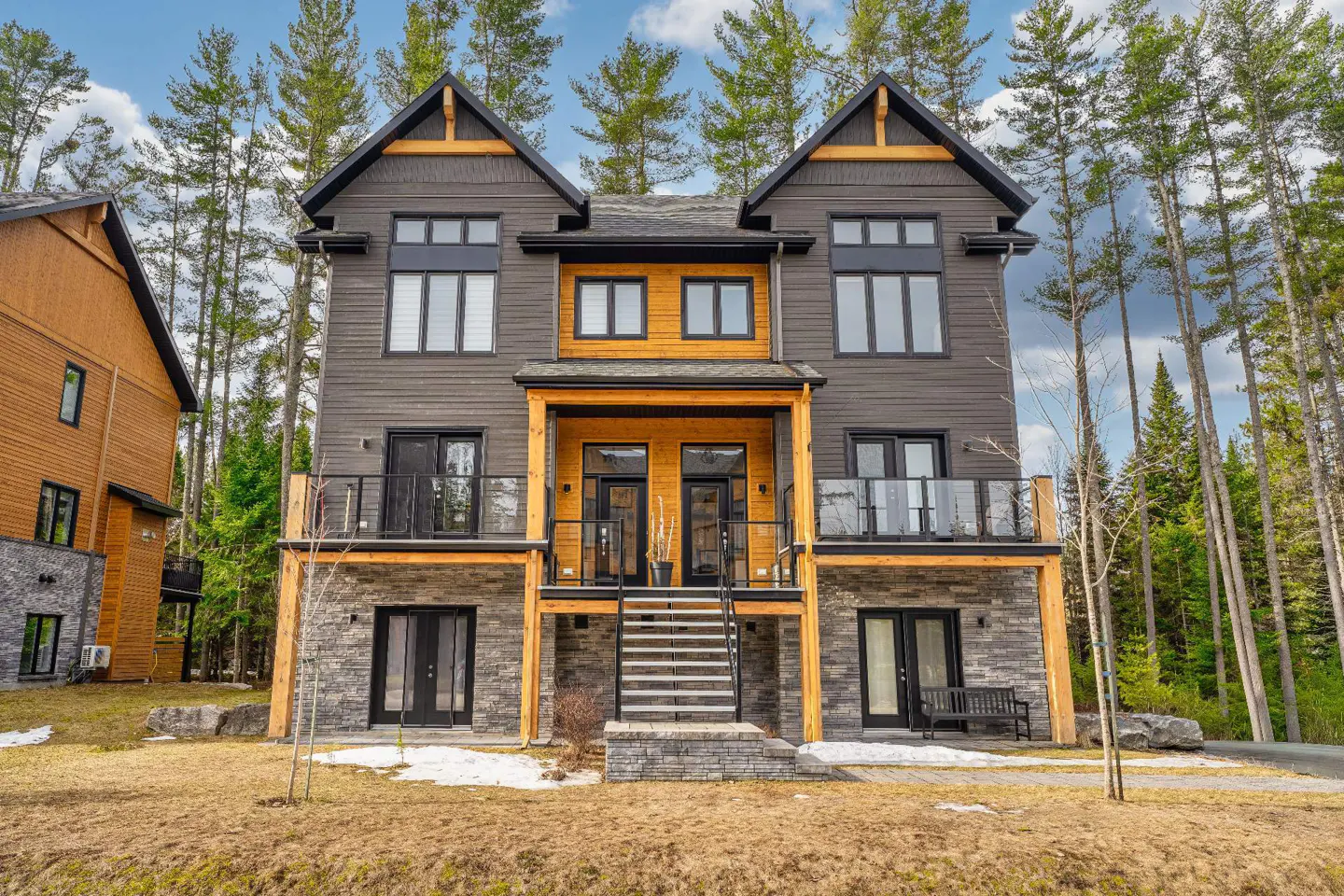 Two-story duplex with gray siding, wood accents, and stone foundation, surrounded by tall pine trees.