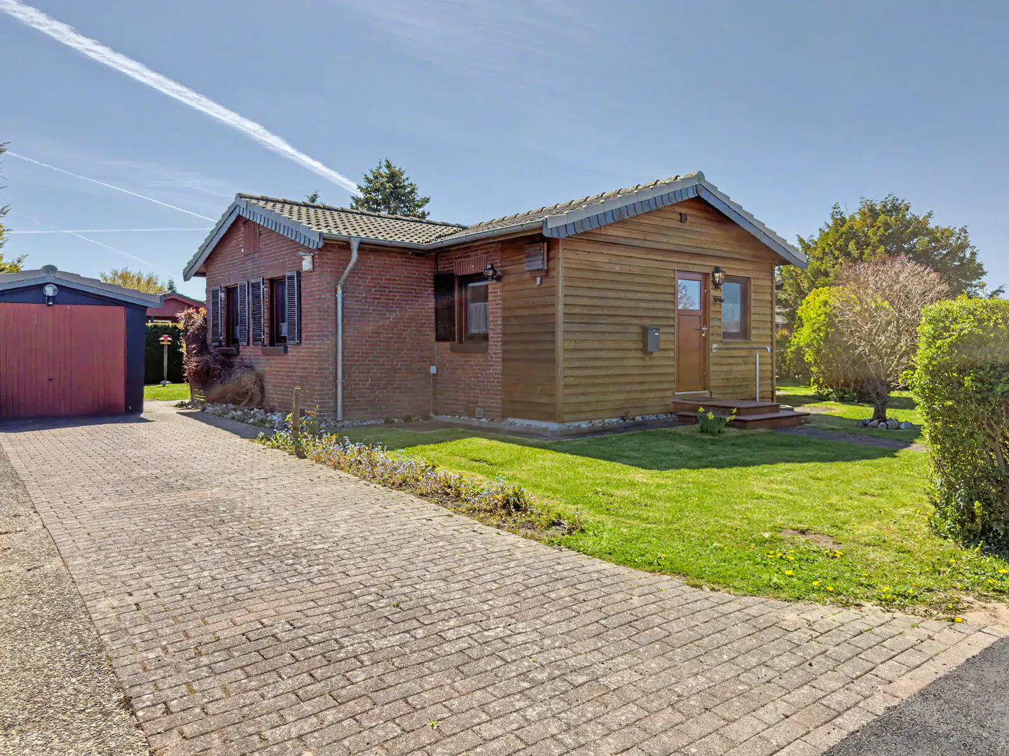 A single-story house with a red brick and wood-paneled exterior, a paved driveway, and a detached garage on a sunny day.