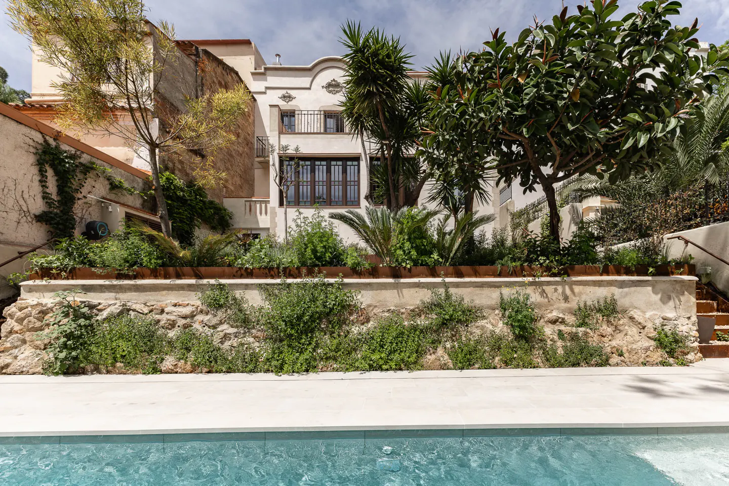 Exterior view of a white building with brown windows, surrounded by lush greenery and a blue swimming pool.