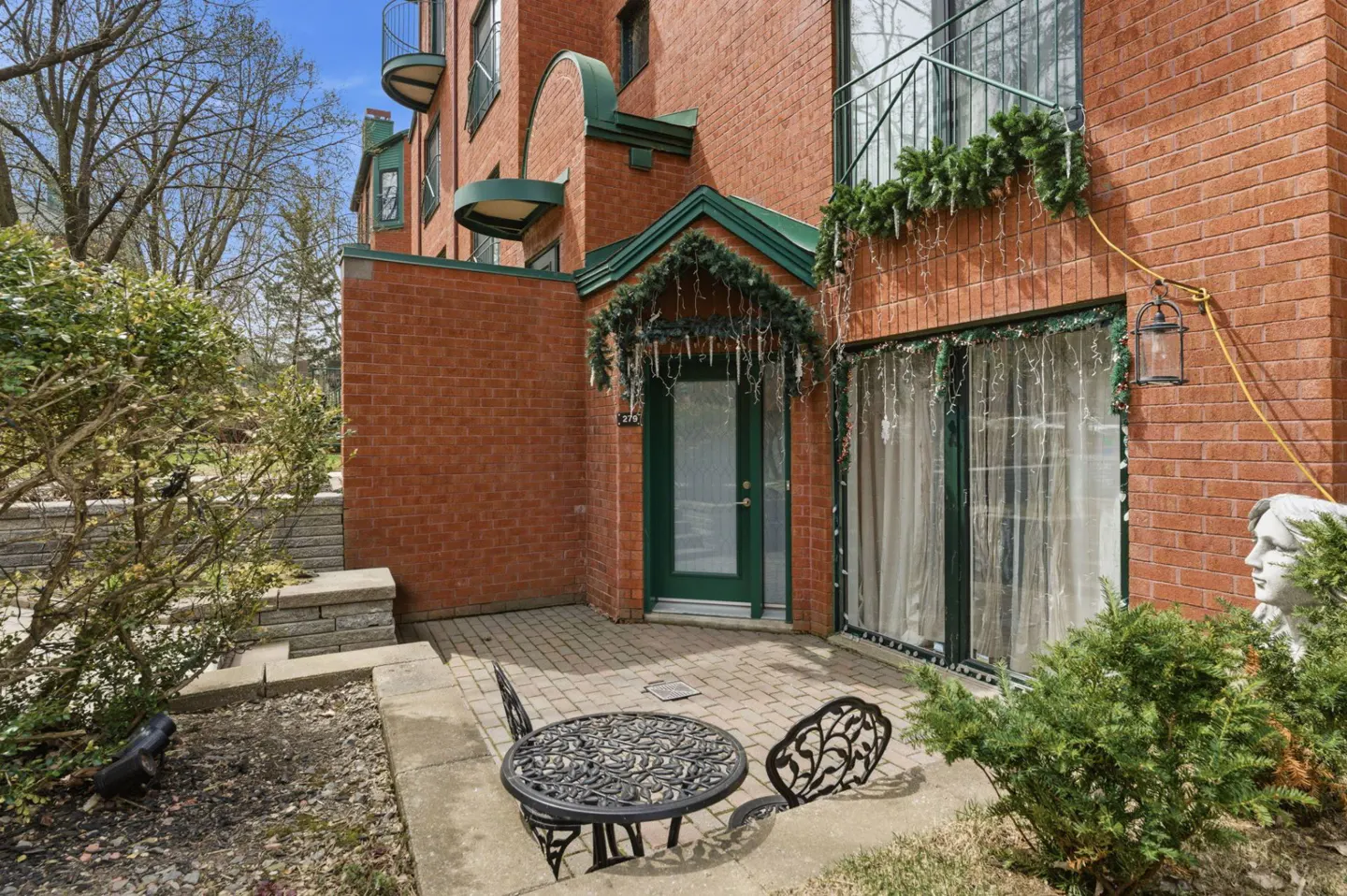 Brick building with a green door and trim, decorated with garland. A patio with a black table and chairs sits outside.