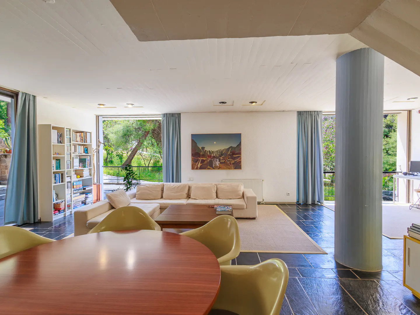 Open-plan living room with a round wood table, olive chairs, and a white sofa. Large windows offer views of lush greenery.