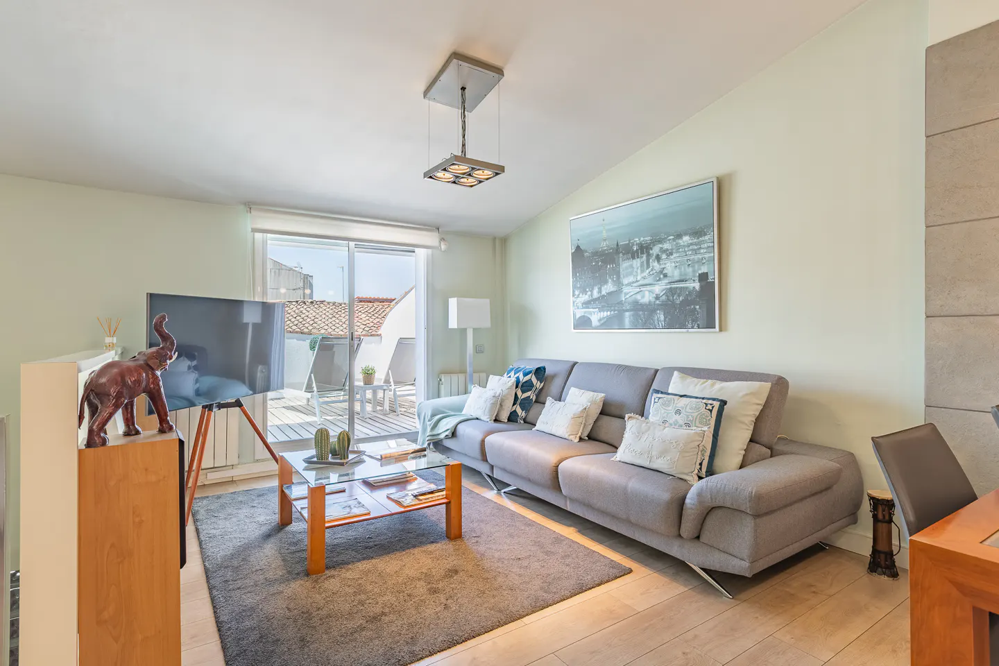 Living room with gray sofa, glass table, and TV on a tripod stand. Balcony access with outdoor furniture. Light green walls and wood floors.