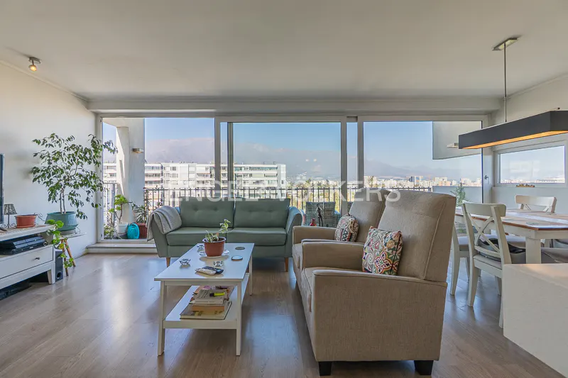 Living room with a green sofa, beige armchairs, and a white coffee table. Large windows offer city views. Dining area with white table and chairs.