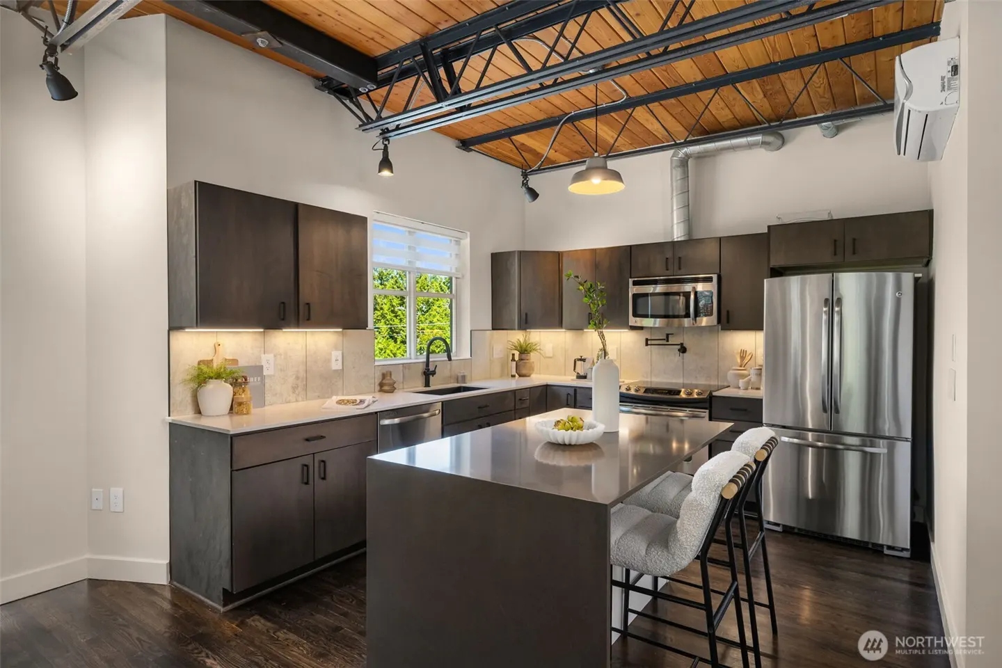 A modern kitchen with dark cabinets, stainless steel appliances, and a large island with seating. The ceiling is exposed wood with metal beams.