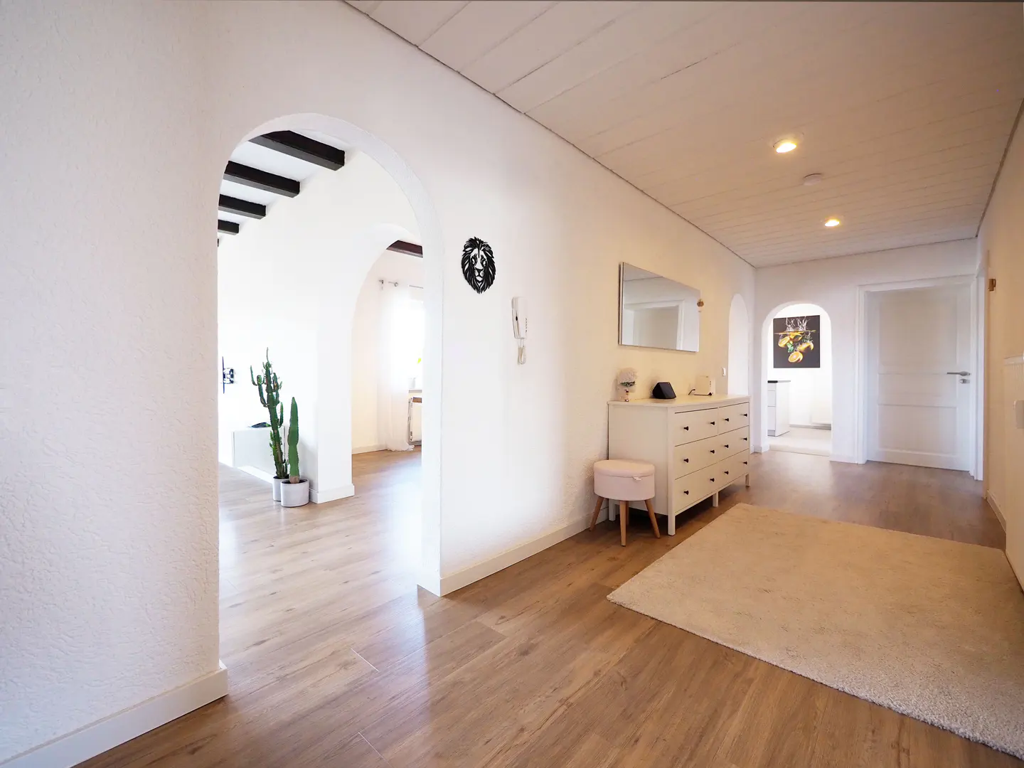 Hallway with white walls, wood floors, and arched doorways. A white dresser, mirror, and rug are visible. A lion head decoration hangs on the wall.