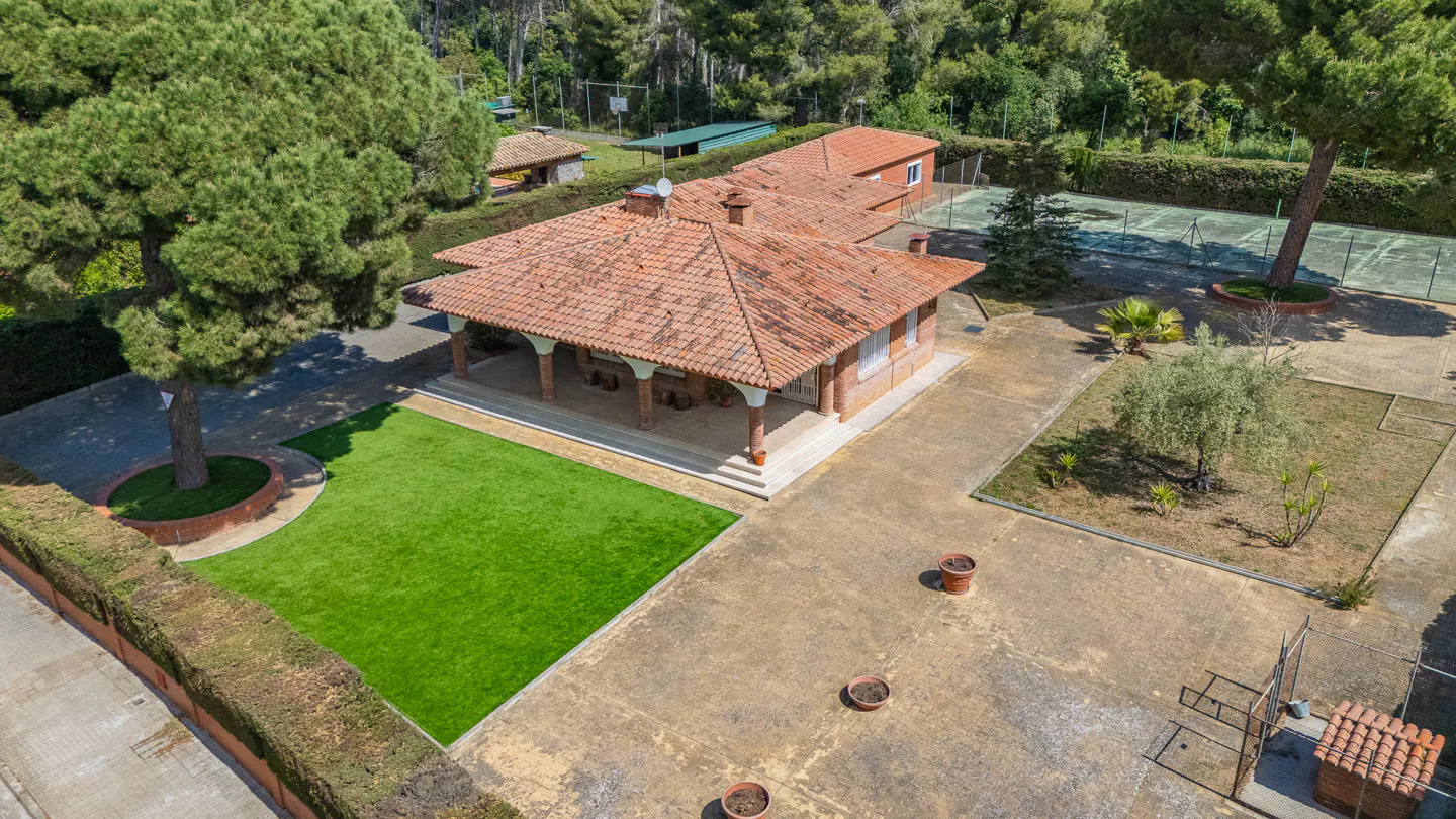 Aerial view of a property with a red-tiled roof house, green lawn, trees, and a tennis court in the background.