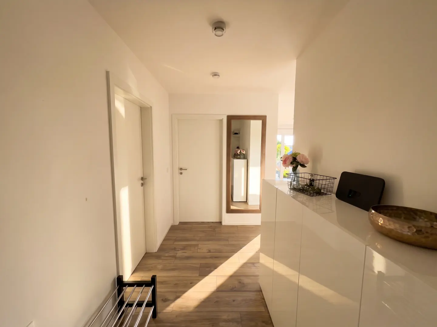 Bright hallway with wood floors, white walls, and doors. A shoe rack is on the left, and a white cabinet with decor is on the right. A mirror hangs on the back wall.