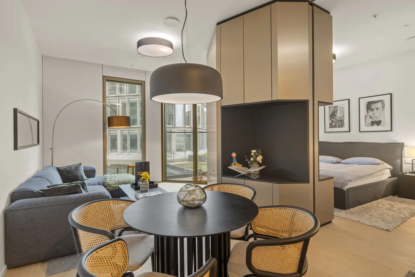 Modern studio apartment with a round black dining table, rattan chairs, grey sofa, and a bedroom area behind a partition.