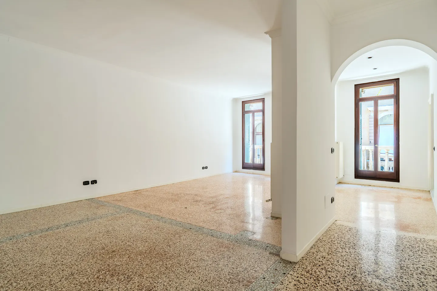 Empty room with white walls, terrazzo flooring, and two arched doorways leading to windows with dark wood frames.