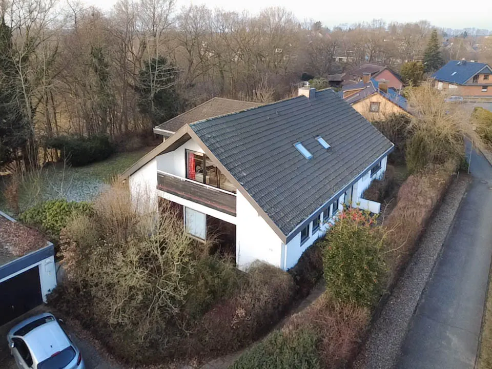 Aerial view of a white, two-story house with a gray roof and a balcony, surrounded by trees and bushes. A car is parked in the driveway.