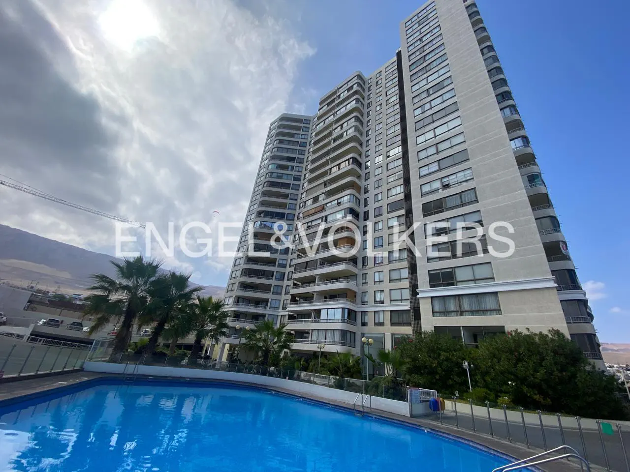 Exterior view of a tall, modern apartment building with a blue swimming pool in the foreground and palm trees. The sky is partly cloudy.