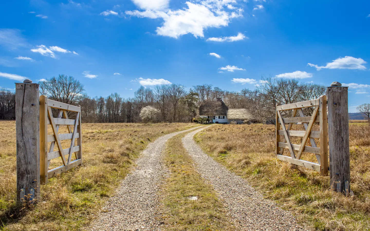 Open wooden gate leads to a gravel path, ending at a white cottage with a thatched roof under a blue sky.