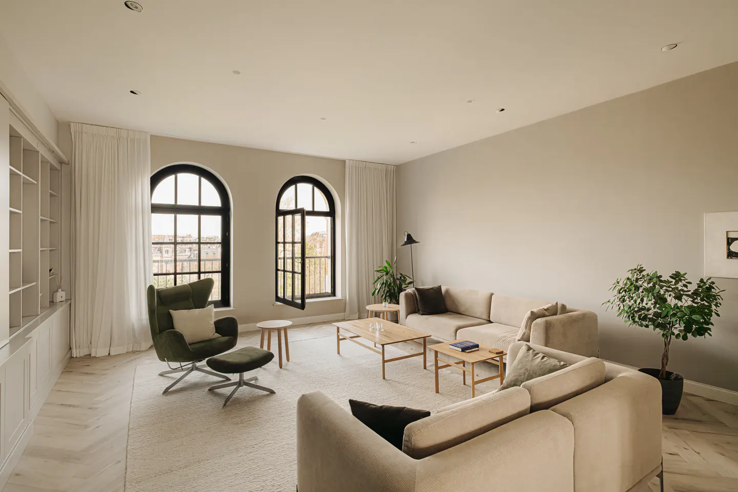 Spacious living room with beige walls, light wood floors, and a large area rug. Two beige sofas, a green armchair, and arched windows with black frames.