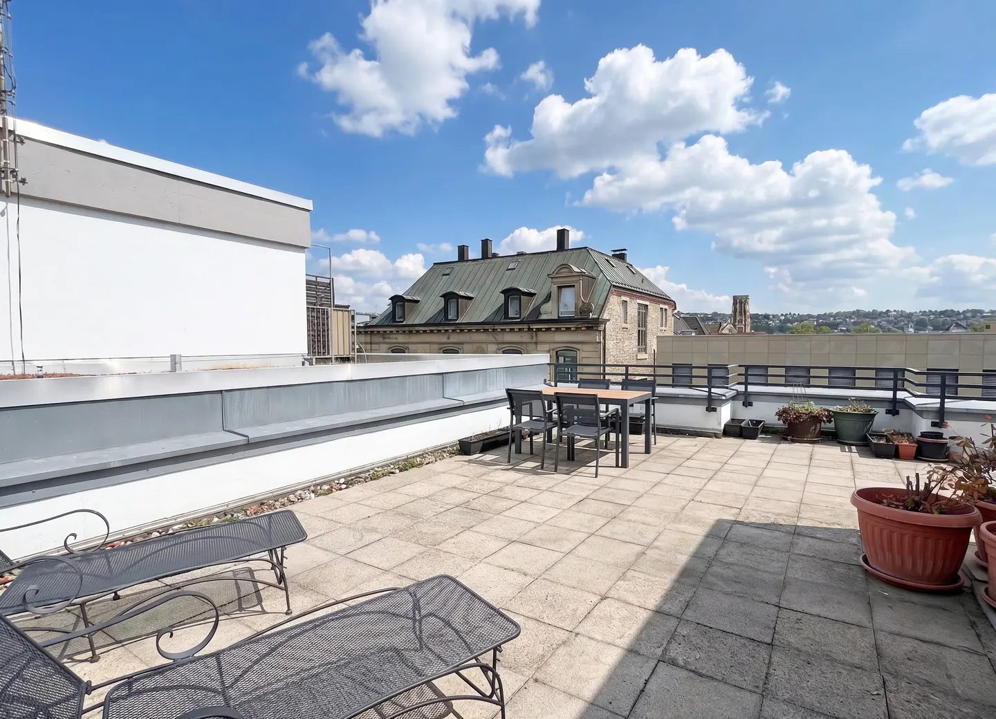 Rooftop patio with lounge chairs, table, and potted plants. A building is visible in the background under a blue sky with white clouds.