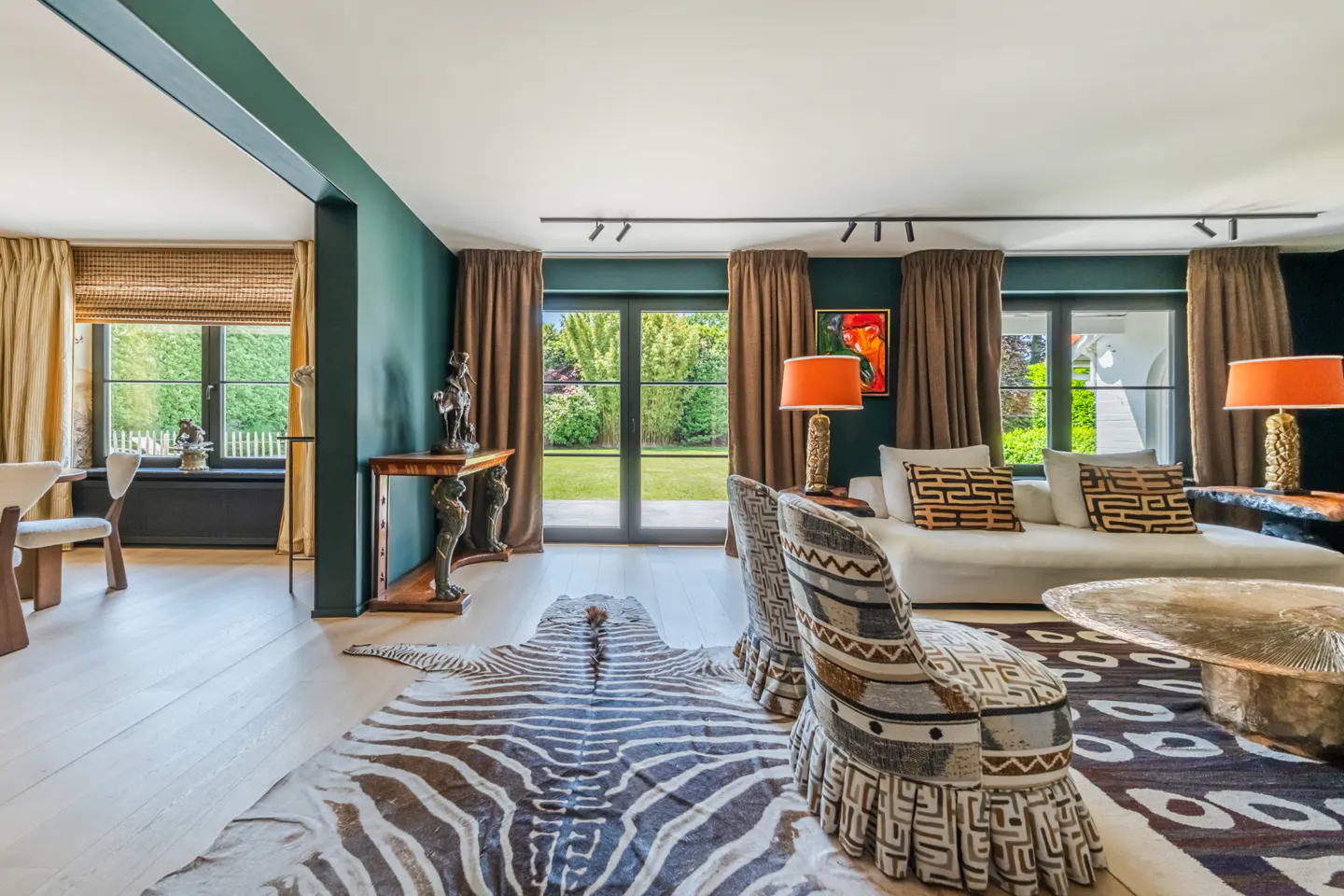 A living room with dark green walls, light wood floors, and a zebra-print rug. A white sofa and patterned chairs are visible.