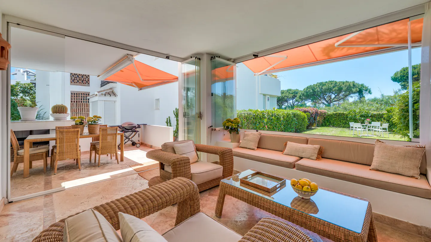 A sunroom with wicker furniture, a glass table with lemons, and a view of a green lawn and trees. Orange awnings provide shade.